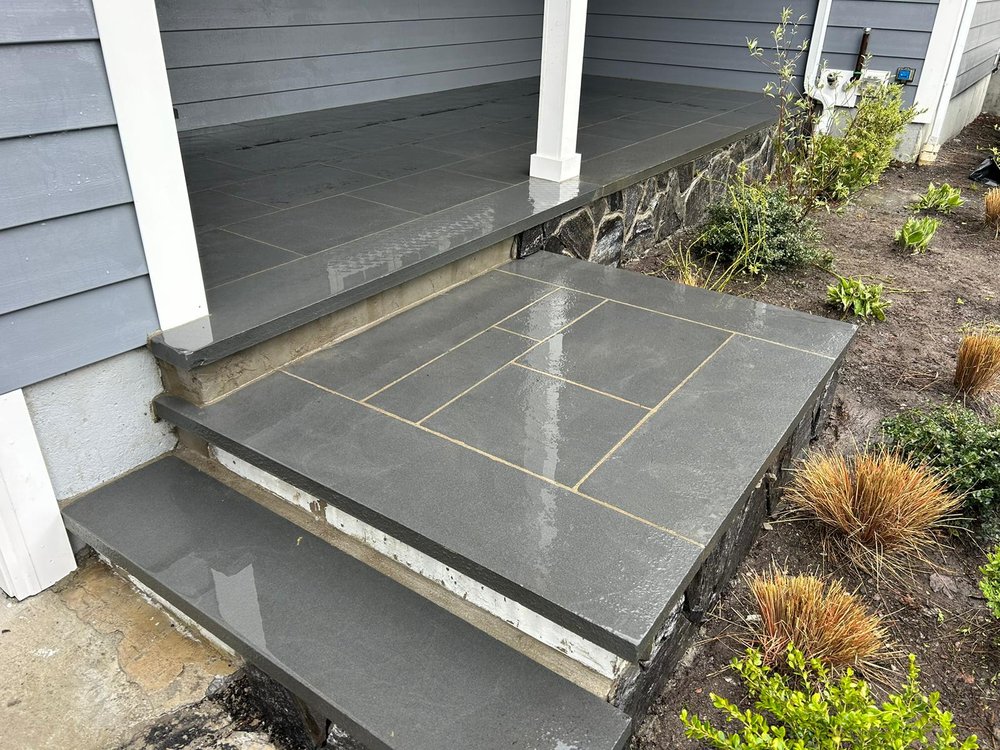 A house entryway with slate tiles, including two tiled steps leading to a porch with similarly tiled flooring. The porch has white pillars and blue-painted walls. Surrounding the entryway are plants and a small garden area with mulch, showcasing the meticulous craftsmanship of Stone Masonry Boston.