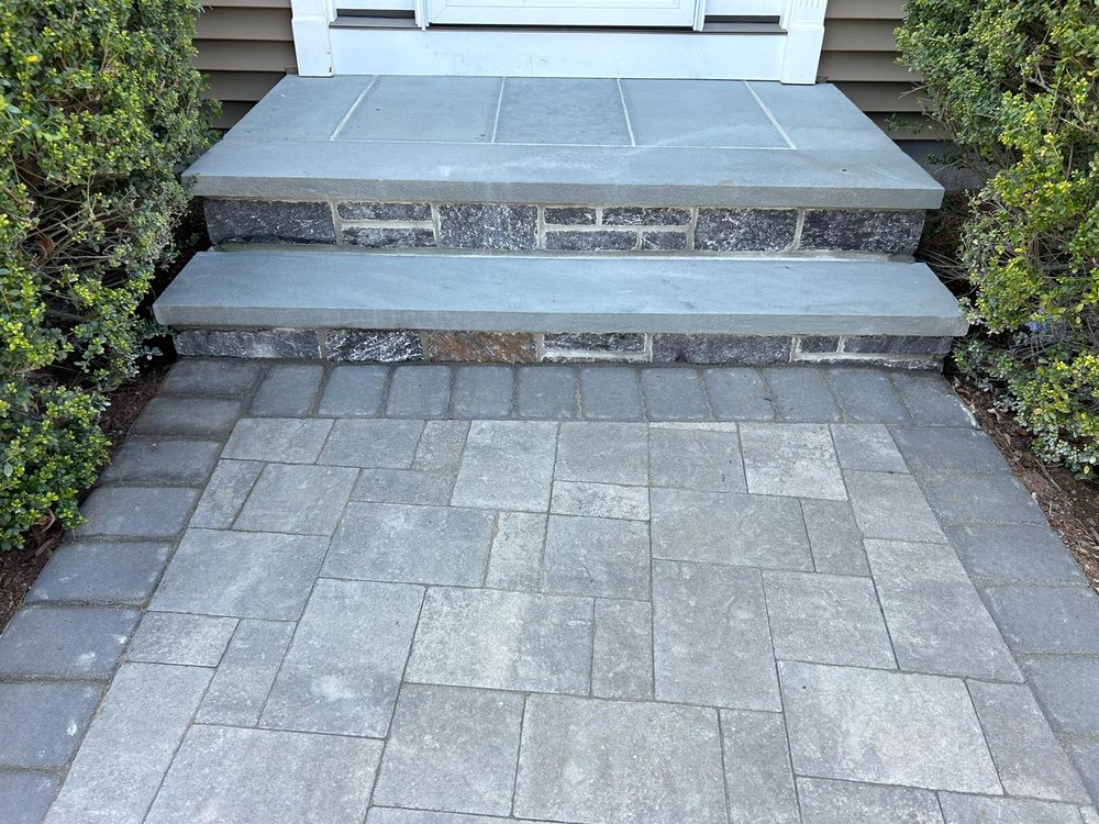 A stone pathway leads to a two-step entrance with a stone facade and bluish-grey stone tops, showcasing impressive Stone Masonry Boston craftsmanship. Green bushes adorn both sides of the steps, and a white door is visible at the top of the entrance. The house's light brown siding complements its elegant design.