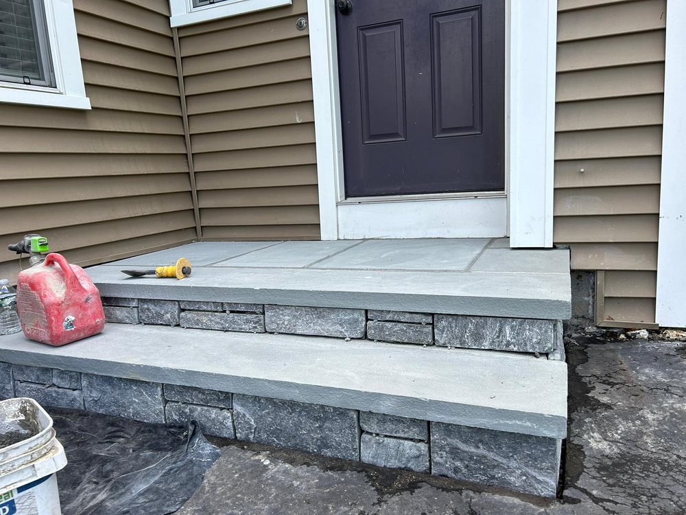 A newly constructed stone staircase leads to a dark-colored door at the entrance of a house with brown siding. Construction tools, including a red fuel canister, chisel, and a plastic sheet, are scattered nearby.